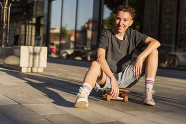 Lächelnde nicht-binäre Person auf Skateboard sitzend - OSF00874