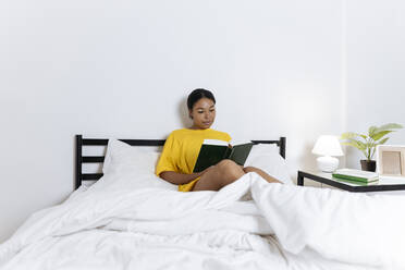 Frau in gelbem T-Shirt sitzt auf dem Bett und liest ein Buch - SIF00393