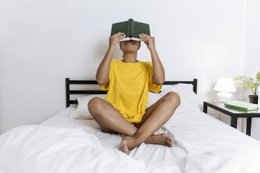 Woman covering her face with a book sitting cross-legged on bed - SIF00396