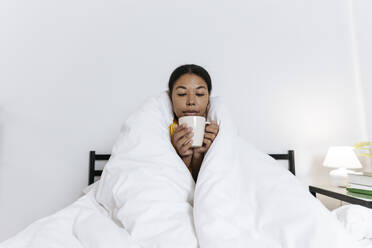 Woman sitting on the bed wrapped in a blanket drinking tea - SIF00415