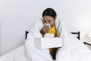 Kranke Frau sitzt auf einem Bett, eingewickelt in eine Decke, und schnäuzt sich mit einem Taschentuch - SIF00416