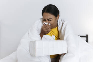 Kranke Frau sitzt auf einem Bett, eingewickelt in eine Decke, und schnäuzt sich mit einem Taschentuch - SIF00417
