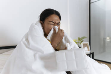 Kranke Frau sitzt auf einem Bett, eingewickelt in eine Decke, und schnäuzt sich mit einem Taschentuch - SIF00418