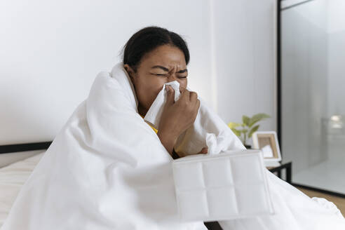 Sick woman sitting on bed wrapped in a blanket blowing nose in a tissue - SIF00418