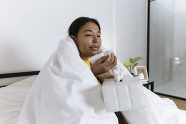 Kranke Frau sitzt auf einem Bett, eingewickelt in eine Decke, und schnäuzt sich mit einem Taschentuch - SIF00419