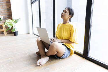 Young woman sitting on floor with eyes closed leaning against a window with computer on lap - SIF00455