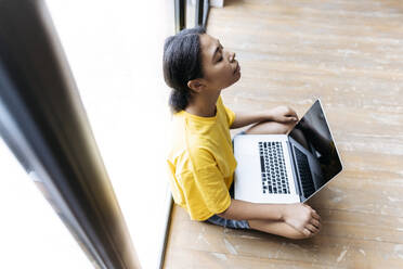 Junge Frau sitzt mit geschlossenen Augen auf dem Boden und lehnt sich mit einem Computer auf dem Schoß gegen ein Fenster - SIF00456