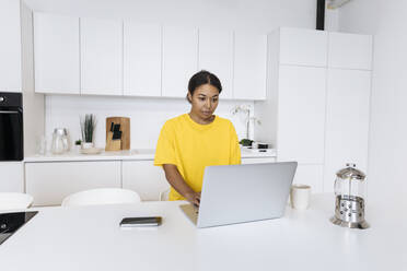 Junge Frau sitzt in der Küche und arbeitet am Laptop - SIF00460