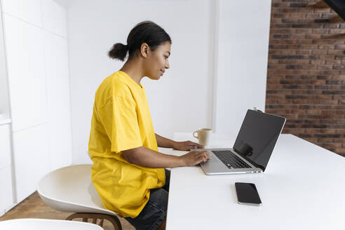 Junge Frau sitzt in der Küche und arbeitet am Laptop - SIF00464