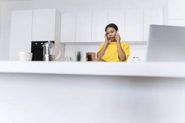 Junge Frau sitzt in der Küche mit Laptop und telefoniert - SIF00467