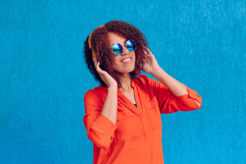 Lächelnde Frau mit Sonnenbrille hört Musik über Kopfhörer vor einer blauen Wand - YTF00001