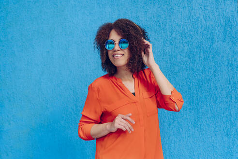 Frau mit Hand im Haar und Sonnenbrille vor einer blauen Wand stehend - YTF00002