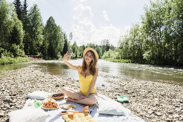 Lächelnde Frau macht ein Selfie am Flussufer und genießt ein Picknick an einem sonnigen Tag - VBUF00153