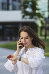 Geschäftsfrau, die die Zeit auf einer intelligenten Uhr abliest und mit einem Mobiltelefon spricht - IHF01170