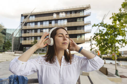 Lächelnde Frau, die vor einem Gebäude Musik über drahtlose Kopfhörer hört - IHF01174