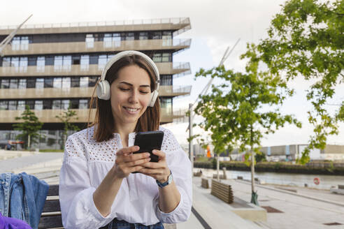 Glückliche Frau hört Musik über kabellose Kopfhörer und benutzt ein Smartphone auf einer Bank - IHF01176