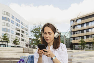 Lächelnde Frau hört Musik über kabellose Kopfhörer und benutzt ein Smartphone auf einer Bank - IHF01177