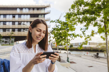 Smiling woman using smart phone in front of building - IHF01179
