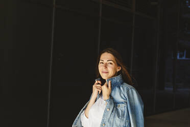 Smiling woman with denim jacket in front of black wall - IHF01186