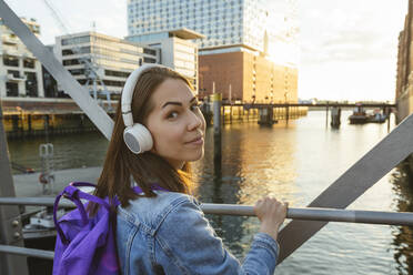 Lächelnde Frau mit drahtlosen Kopfhörern auf der Brücke vor der Elbphilharmonie, Hafencity, Hamburg, Deutschland - IHF01194