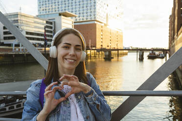Happy woman with wireless headphones gesturing heart shape on bridge in front of Elbphilharmonie, Hafencity, Hamburg, Germany - IHF01195