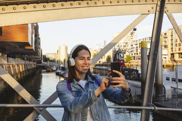 Glückliche Frau mit kabellosen Kopfhörern macht ein Selfie mit ihrem Smartphone auf einer Brücke in der Hafencity, Hamburg, Deutschland - IHF01196