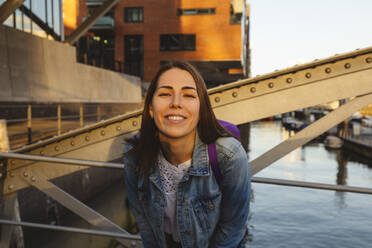 Lächelnde Frau mit Rucksack auf Brücke über Fluss, Hafencity, Hamburg, Deutschland - IHF01197