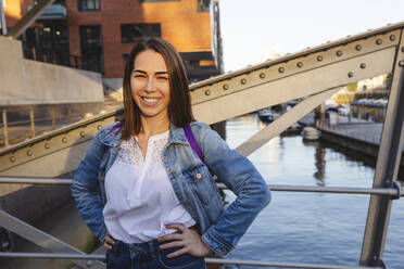 Happy woman with hands on hip standing on bridge over river, Hafencity, Hamburg, Germany - IHF01198