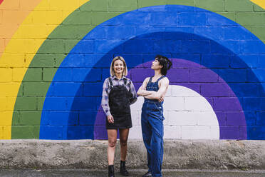 Happy young woman standing with friend in front of rainbow painted on wall - AMWF00824