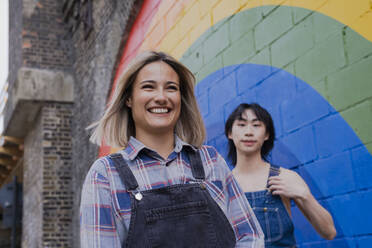 Happy young woman walking with friend by rainbow painted on wall - AMWF00829