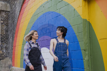 Cheerful young woman and man walking together by rainbow mural on wall - AMWF00830