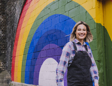 Glückliche junge Frau, die an einer Regenbogen-Wandmalerei vorbeigeht - AMWF00831