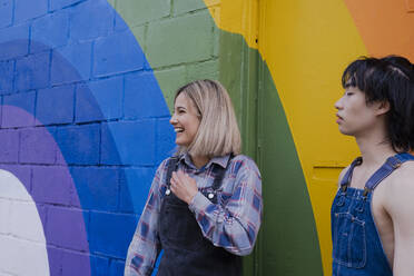 Glückliche junge Frau steht mit ihrem Freund vor einem Regenbogen-Wandbild - AMWF00833