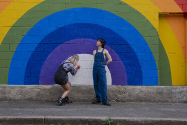 Young man wearing overalls standing by friend writing on rainbow mural - AMWF00836