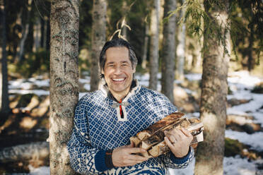 Portrait of happy mature man with firewood standing by tree in forest - MASF31441