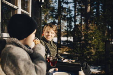 Lächelnder Junge im Gespräch mit einem Freund bei einem Drink auf der Veranda im Winter - MASF31444