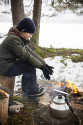 Seitenansicht eines Jungen, der sich die Hände wärmt, während er im Winter am Lagerfeuer sitzt - MASF31471