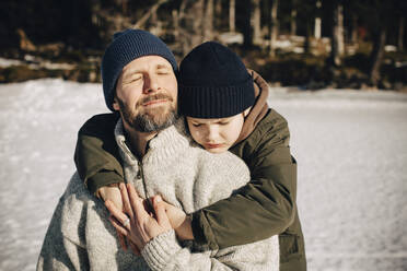 Boy in warm clothing hugging father with eyes closed during sunny day in winter - MASF31529