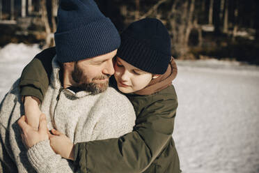 Affectionate father and son wearing knit hat embracing each other on sunny day - MASF31530