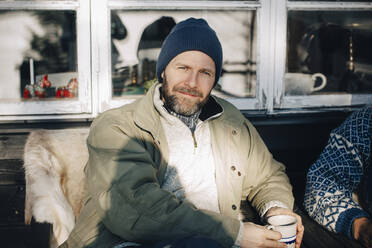 Porträt eines lächelnden reifen Mannes, der mit einer Kaffeetasse auf einer Veranda im Winter sitzt - MASF31533