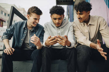 Junge Männer sehen einen Freund an, der ein Smartphone benutzt, während sie im Freien sitzen - MASF31697