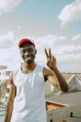 Porträt eines jungen Mannes mit Mütze, der das Friedenszeichen gegen den Himmel an einem sonnigen Tag gestikuliert - MASF31717