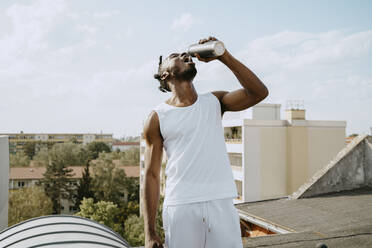 Young man drinking water from bottle while standing on rooftop during sunny day - MASF31721