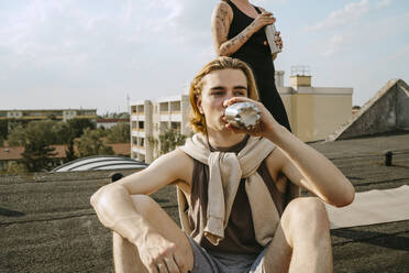 Junger Mann trinkt Wasser, während er vor einem Freund auf dem Dach sitzt - MASF31740