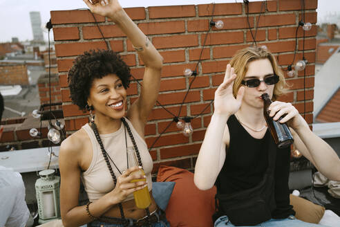 Glückliche Frau und Mann mit erhobenen Händen beim Biertrinken auf einer Dachterrasse - MASF31752