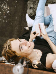 Portrait of smiling young man with book sitting on rooftop - MASF31763