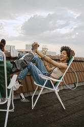 Full length of cheerful woman holding hand of male friend sitting on chair at rooftop - MASF31769