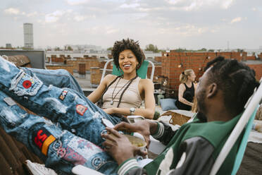 Happy young woman looking at friend sitting on chair at rooftop - MASF31770