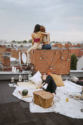 Frau auf Decke liegend mit Freundinnen, die auf einem Dach sitzen und sich umarmen - MASF31788