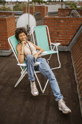 Full length portrait of smiling young woman sitting on chair at rooftop - MASF31798